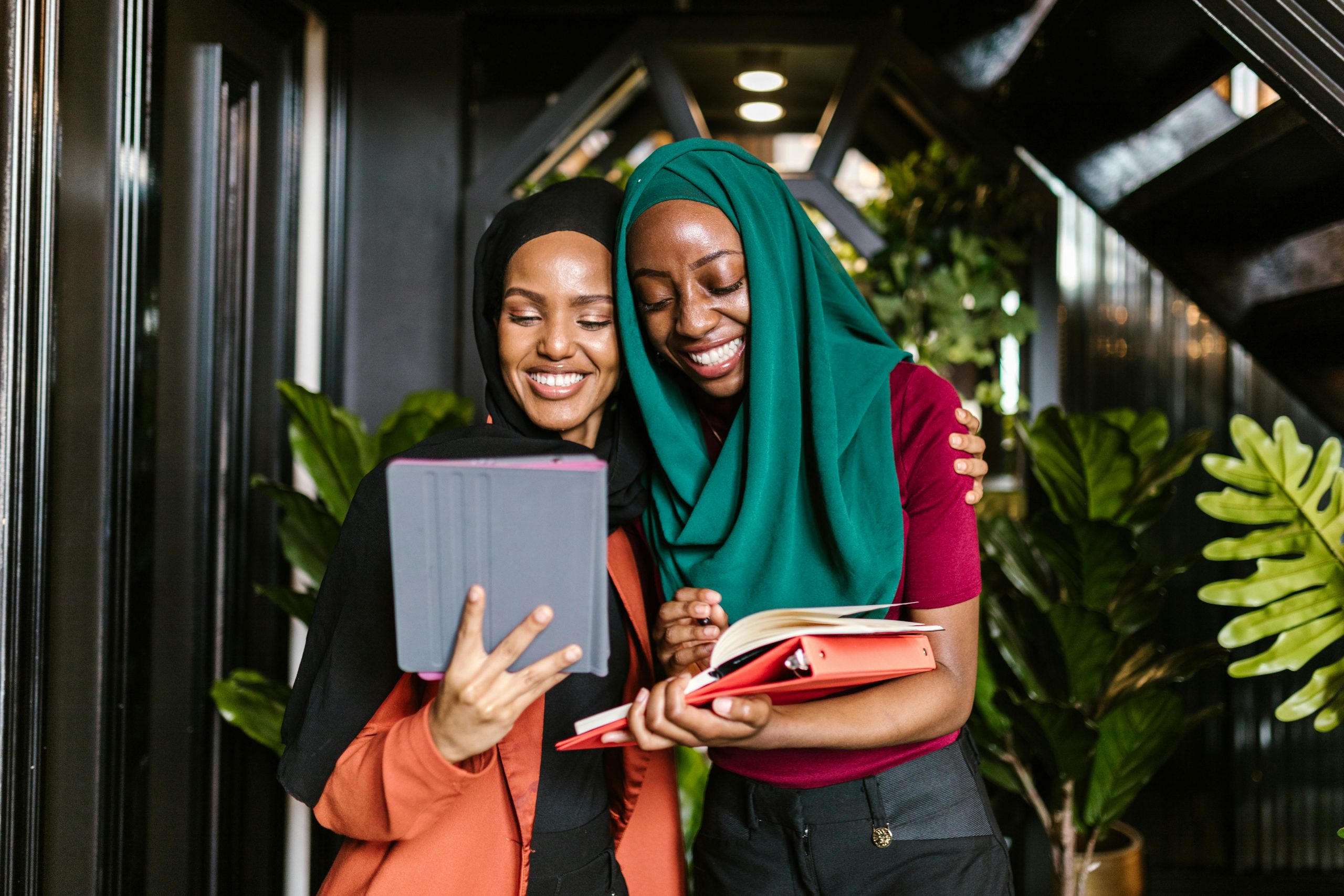 Two women in hijabs smiling together, using an iPad and notebook in an office setting.